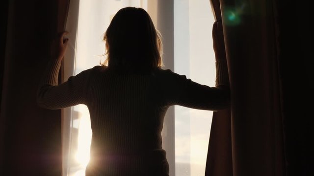 The silhouette of a woman who opens the curtains on the window and looks forward