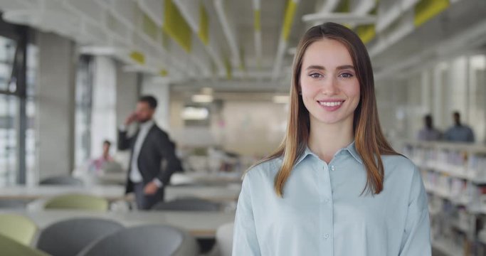 Young pretty female office worker turning head and looking to camera. Portrait of happy millennial female employee smiling and standing in office workroom. Concept of people and workspace.