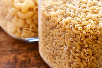 Closeup of uncooked elbow macaroni and pipe rigate in glass jars on a wooden table. Image with selective focus.  