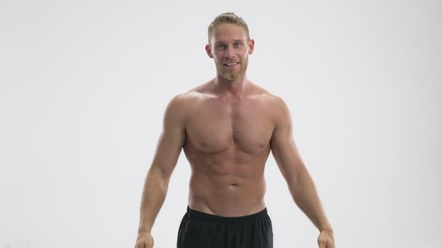 A strong young shirtless sportsman is showing his biceps while raising his hands isolated over white background