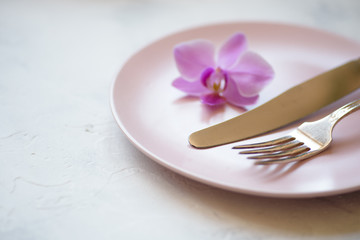 Steel fork and knife with a purple orchid flower on a pink plate on white background