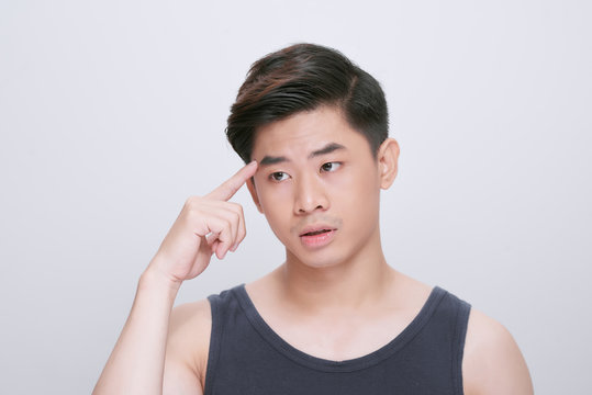 Young asian chinese man wearing t-shirt standing over isolated white background pointing unhappy to pimple on forehead, ugly infection of blackhead. Acne and skin problem