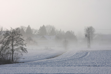 Fototapeta premium Electricity pylon at the edge of the village in winter, snowy foggy rural landscape in the morning light