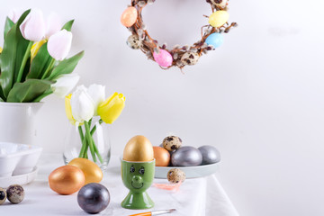 Holiday composition of served table with golden egg in a green cup as a face , baked cookies, fresh spring tulips flowers and festive wreath on a light grey wall background. Happy Easter concept.
