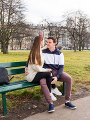 Young couple sitting on a wooden bench. Beautiful sunny day. Young happy people relaxing outdoor in the park. They are looking at each other. Travelers