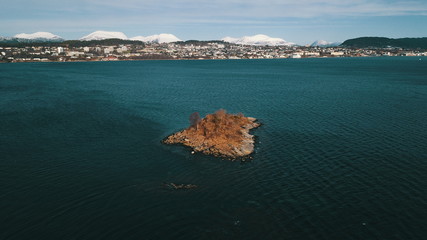 A small, rocky island, not far from the shore. Behind the city is the backdrop of snowy mountains.