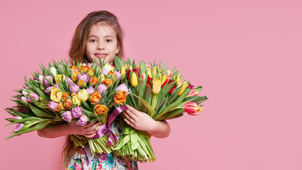 Cute smiling child girl holding bouquet of spring flowers tulips isolated on pink background. Little toddler girl gives a bouquet to mom. Copy space for text.