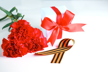 Three red carnation flowers, St. George ribbon, a gift in a white box with a red bow on a white...