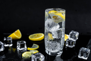 Sparkling water with lemon and ice, white soda in glass on black background, refreshing cold drink