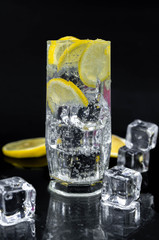 Sparkling water with lemon and ice, white soda in glass on black background, refreshing cold drink