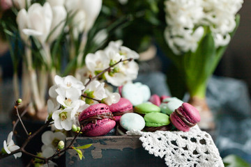 Beautiful Easter composition with bright delicious homemade traditional french dessert - elegant macarons. Floral decor, spring flowers on background, vintage wooden box. Front view, close up.