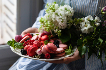 Young beautiful girl in blue shirt is holding in her hands a plate with delicious homemade french macarons - traditional french dessert. Concept of fragile beauty, tenderness, floral wedding decor