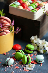 Assortment of bright delicious homemade traditional french macarons in the gift box. Natural fruit and berry flavors, sweet creamy stuffing. Close up, dark background. Fresh spring flowers as decor