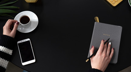 Hand of woman holding cup of coffee and pencil with notebook and phone with blank screen with black background with copy space. Office desk and business with top view. Panoramic banner