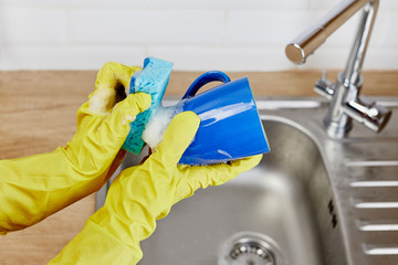 Hands with sponge wash the cup under water, housewife woman in yellow rubber protective gloves washing blue mug