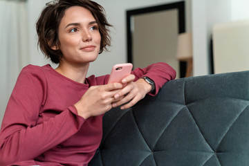 Photo of pleased young woman using smartphone while sitting on sofa