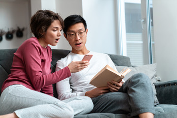 Photo of shocked multinational couple reading book and using smartphone