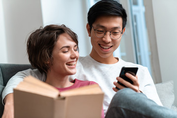 Photo of laughing multinational couple reading book and using cellphone