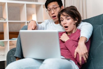 Photo of shocked multinational couple hugging and using laptop