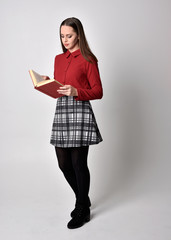 full length portrait of a pretty brunette girl wearing a red shirt and plaid skirt with leggings and boots. Standing pose holding a camera, on a  studio background.