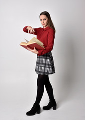full length portrait of a pretty brunette girl wearing a red shirt and plaid skirt with leggings and boots. Standing pose holding a camera, on a  studio background.