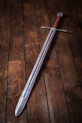 A metal sword on a wooden table with a brown hilt and an unusual guard. Witcher sword made of wood and painted with silver paint. Steel blade.