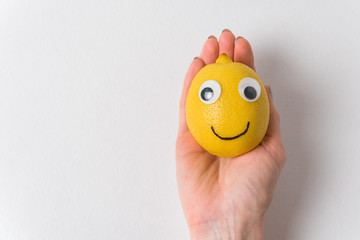 Funny lemon fruit character lies in female hand on white background. Cheerful fruit with eyes and...