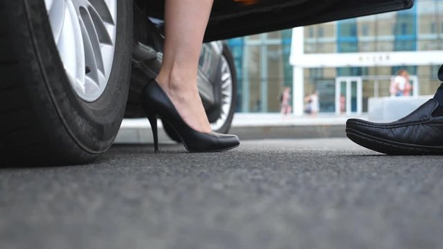 Businessman opens and holds door of car for beautiful young woman. Slim legs of businesswoman in shoes on high heels getting out of auto. Close up Slow motion Low angle view