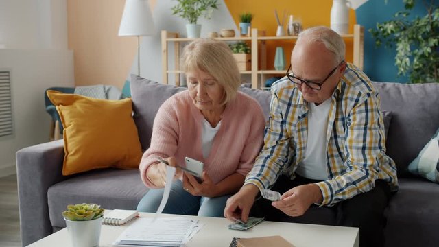 Senior couple is dealing with financial papers and counting money in apartment looking through bills together. Financial documents and cash concept.