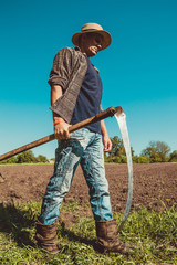 Authentic rural farmer with scythe. Agriculture worker. Farm implements. Rustic background. Work countryside. Brutal country man. Manual labor. European farmland. Agrotourism concept. Vertical