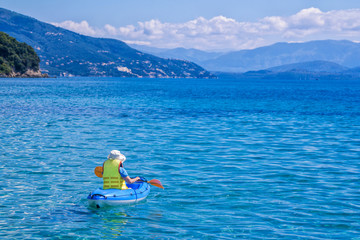 Woman paddling kayak in a calm sea bay with turquoise water and mountains on the horizon. Summer activity, Corfu Island, Greece. 