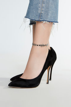 Cropped shot of female legs with an ankle bracelet made as a double row of metal beads on the black cord and inserted silver starfishes. The lady is wearing black high heels and fringed jeans.  