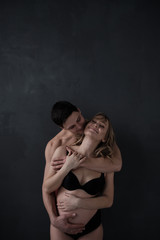 Handsome  man and his beautiful pregnant wife are hugging and smiling while standing near the window at home