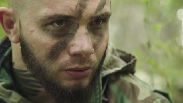 Angry fighter in camouflage with soot strips on face starring in forest