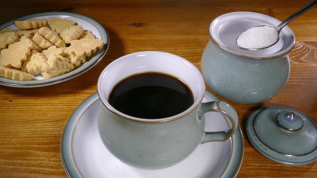 Cinemagraph – Video of steaming black coffee in a cup after having been stirred, with a still image of a spoonful of sugar from a bowl ready to be added, and a plate of biscuits. 