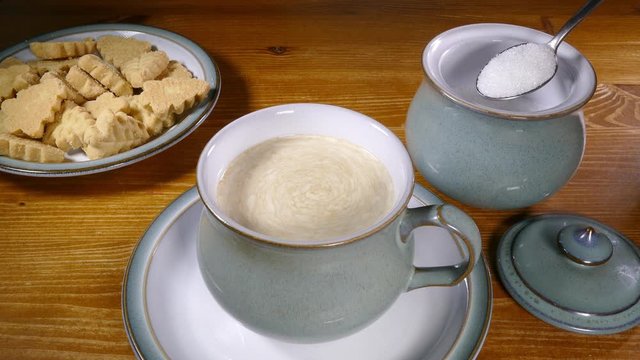 Cinemagraph – Video of cream on top of coffee, swirling in a cup after having been stirred, with a still image of a spoonful of sugar from a bowl ready to be added, and a plate of biscuits. 