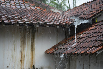 Rain falls from a brown tile roof over a white wall.