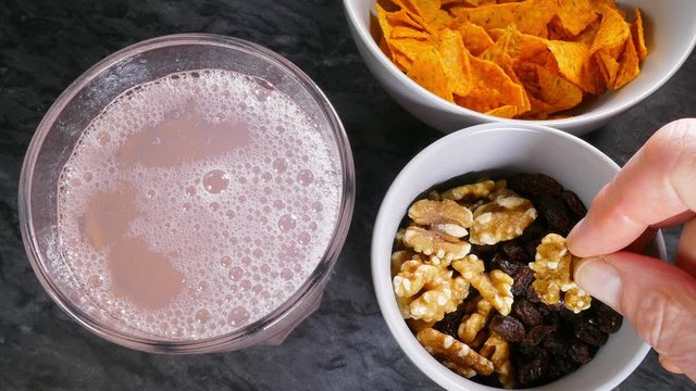 Cinemagraph – Video of fizzing pink grapefruit soda carbonated drink in a glass, with a still image of fingers holding a walnut from a snack bowl also containing raisins, next to tortilla chips. 