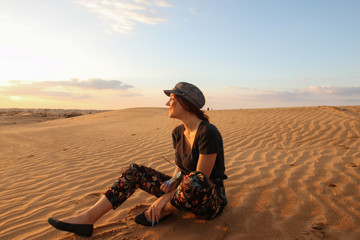 Happy young woman with hat sitting in the desert dunes. Travel, vacations and lifestyle concept.