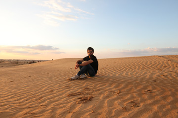 Young hipster man with hat and sunglasses sitting on desert dunes
