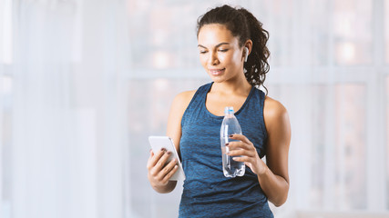 Latin woman with bottle of water, smartphone and earbuds listening to music in gym, blank space