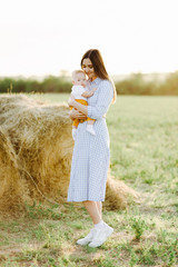 a young pretty mother with a small child in her arms stands in a dress in the middle of a field near a collected haystack in the rays of the Golden, sunset, summer sun