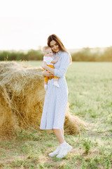 a young pretty mother with a small child in her arms stands in a dress in the middle of a field near a collected haystack in the rays of the Golden, sunset, summer sun