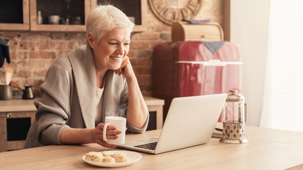 Happy Aged Lady Relaxing In Kitchen With Laptop And Coffee
