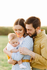 portrait of a happy young family, where mom and dad hug each other, holding their little child between them in the rays of the sunset, summer sun