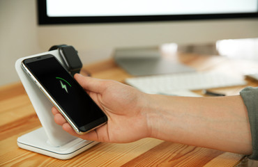 Man taking mobile phone from wireless charger at wooden table, closeup. Modern workplace accessory