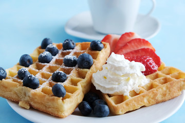 Viennese waffles on a white plate with fresh organic strawberry and blueberry berries and fresh whipped cream and a cup of coffee or tea in the background. Close-up. Blue background. Snack, breakfast.