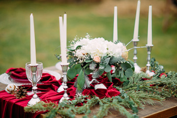 Wedding banquet table with candles, plates, glasses, flower arrangement and a red tablecloth.