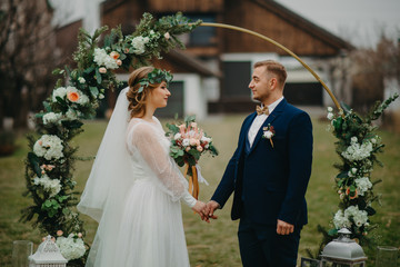 Newlyweds stand near wedding arch and hold hands.
