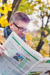 Naklejka premium Portrait of mature attractive man sitting on bench while reading newspaper in park